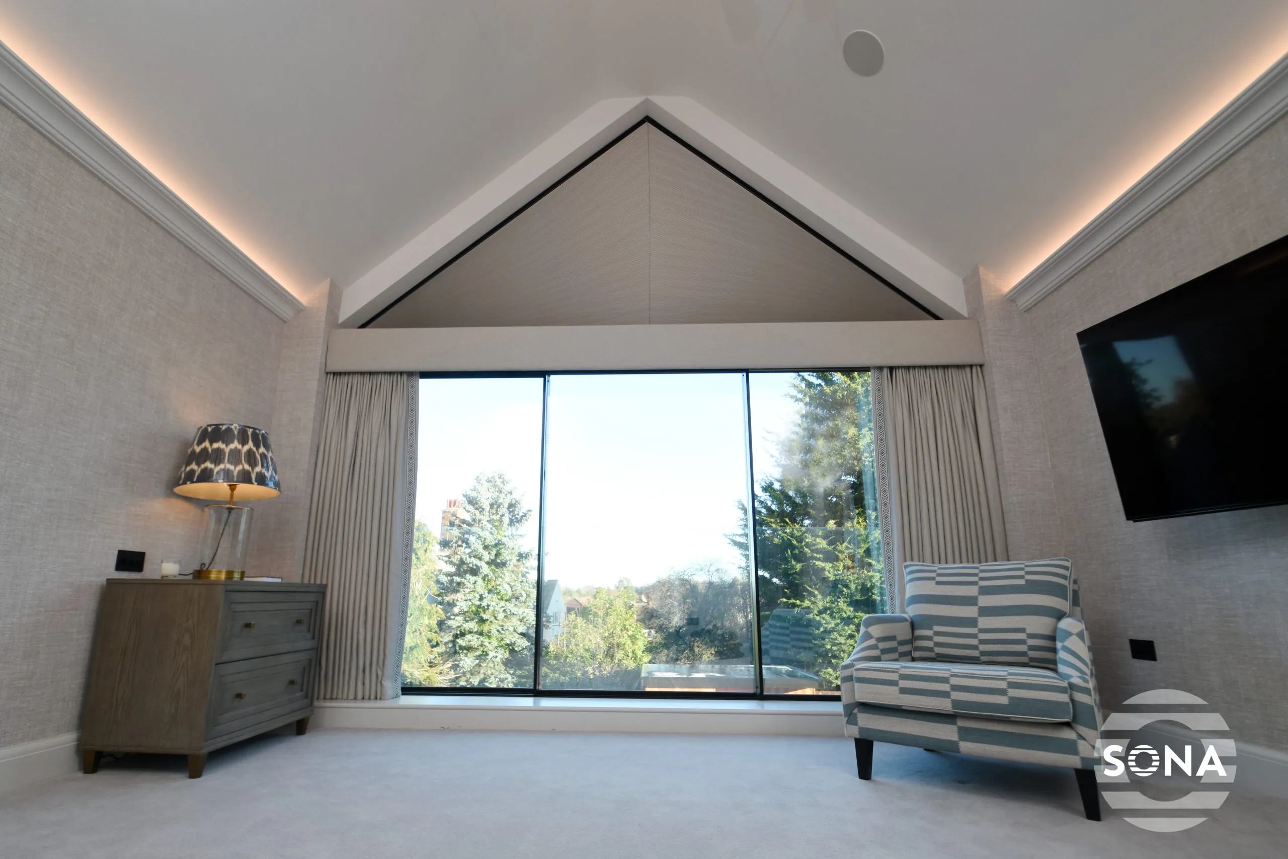 a room with a desk and chair with a large shaped apex window with the top covered by an electric apex window blind.